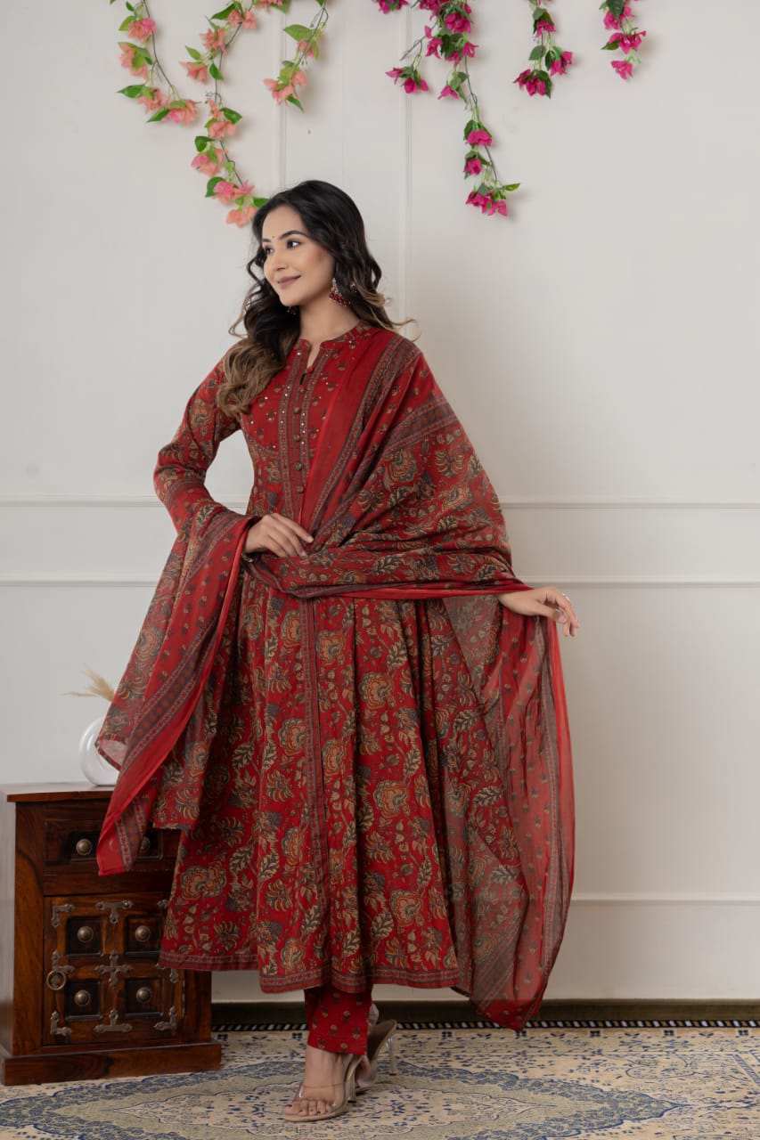 Woman in a red traditional outfit with a patterned shawl against a white wall with floral decorations.